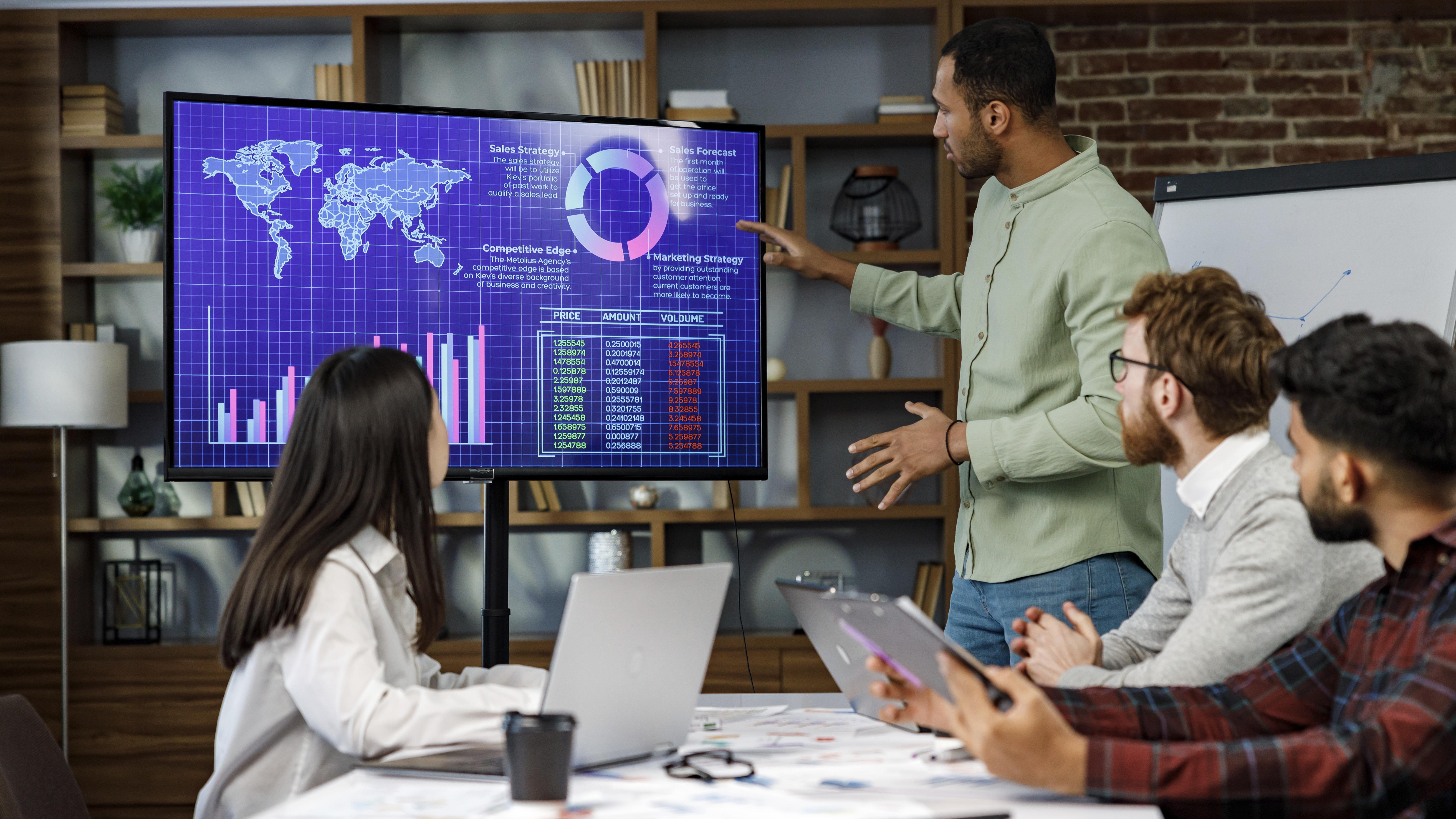 Four adults in a meeting room. A man stands and points at a data monitor while the others work at laptops and tablets on company strategy and data analysis.