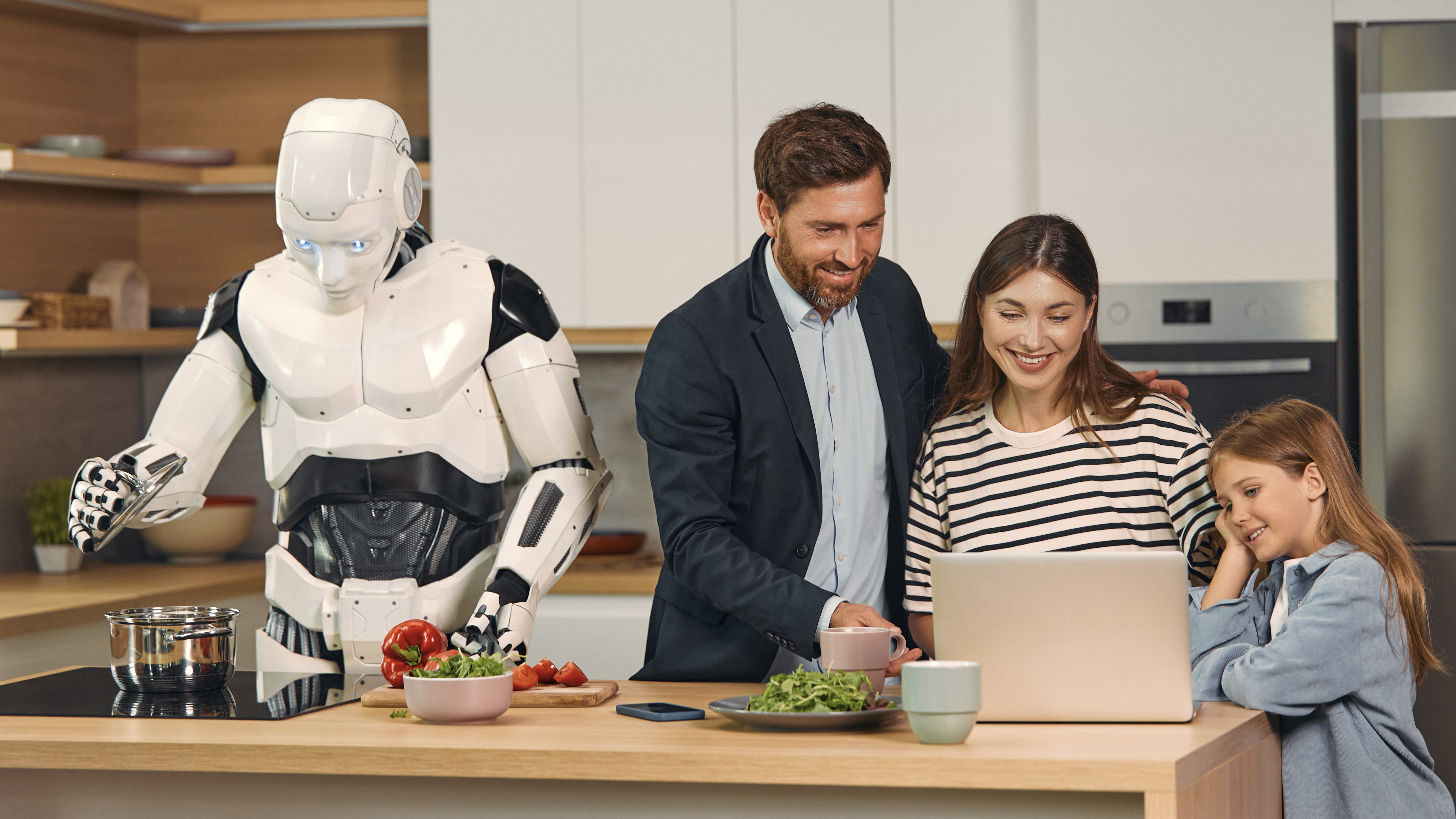 Un homme souriant, une femme et un enfant regardent un ordinateur portable sur un îlot de cuisine, tandis qu'un robot blanc et noir les aide à cuisiner.