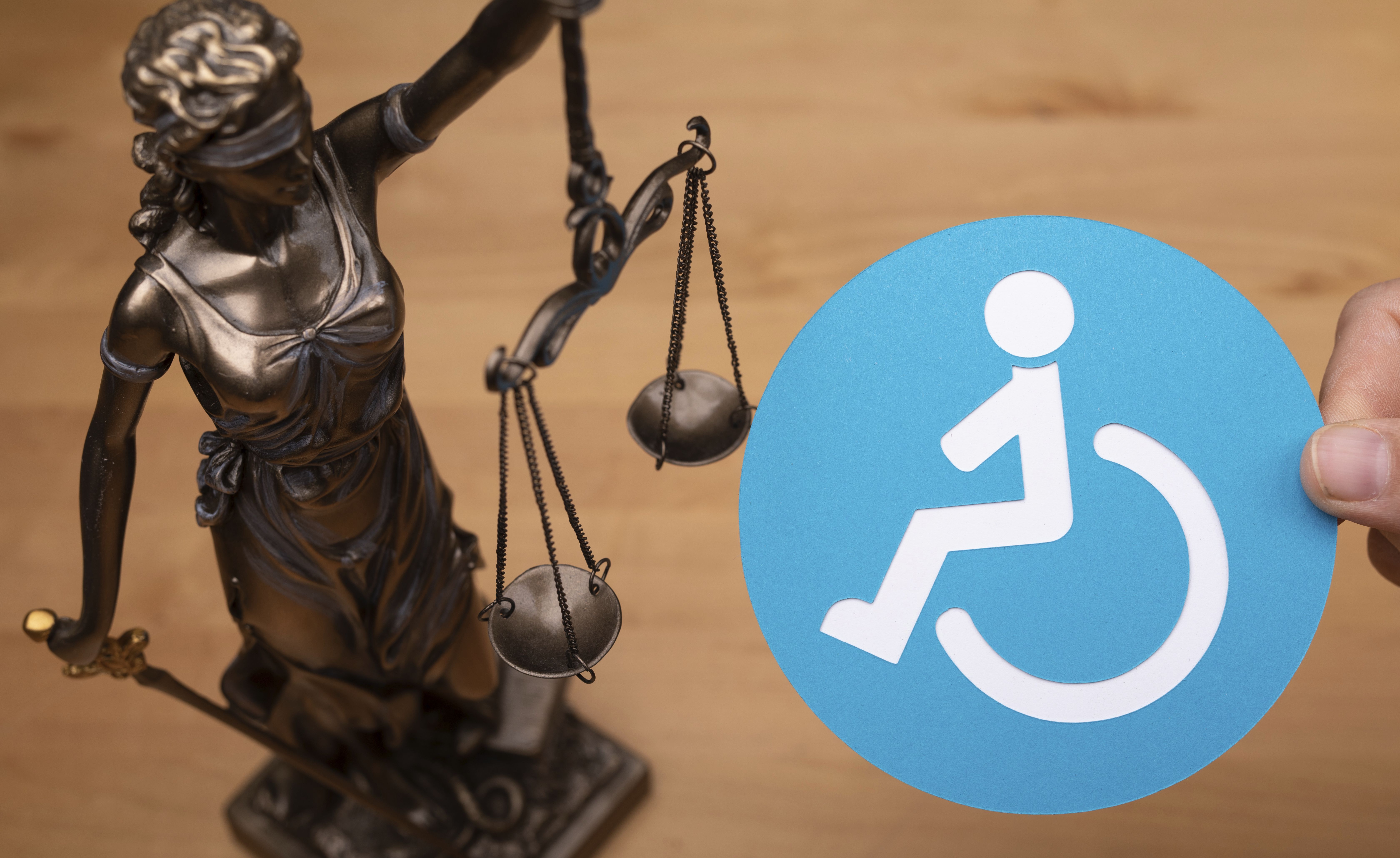 A bronze-colored statue of Justitia stands on a wooden platform next to a blue circle with a white wheelchair symbol. The composition is brightly lit and could symbolize justice, accessibility, and equal rights for people with disabilities.