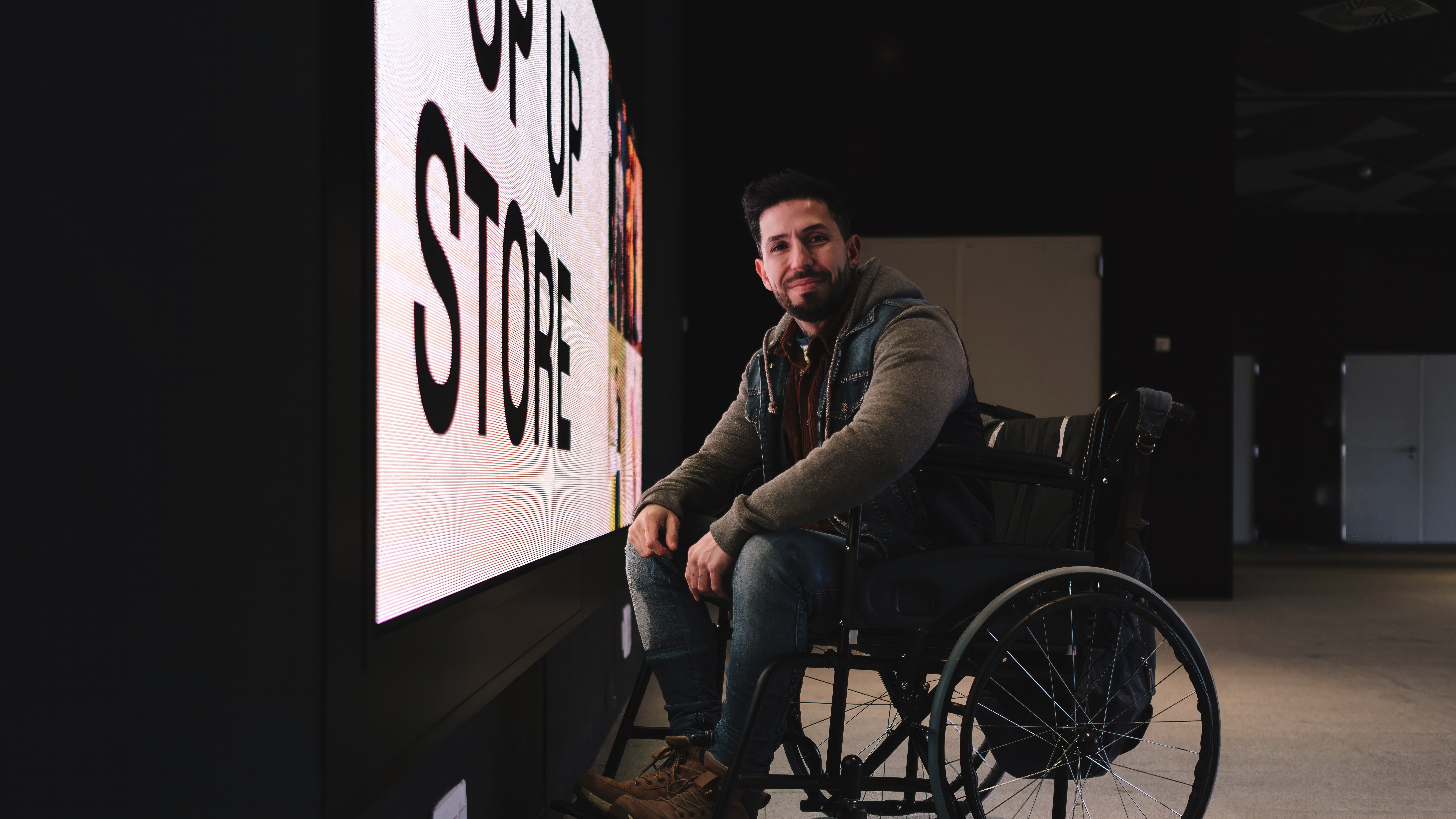 En skægget mand sidder i kørestol foran en sort væg med det lysende skilt »POP UP STORE«. Han har en denimjakke på over en grå hættetrøje, blå jeans og brune sko, og armene hviler løst på armlænene. Han kigger direkte ind i kameraet.