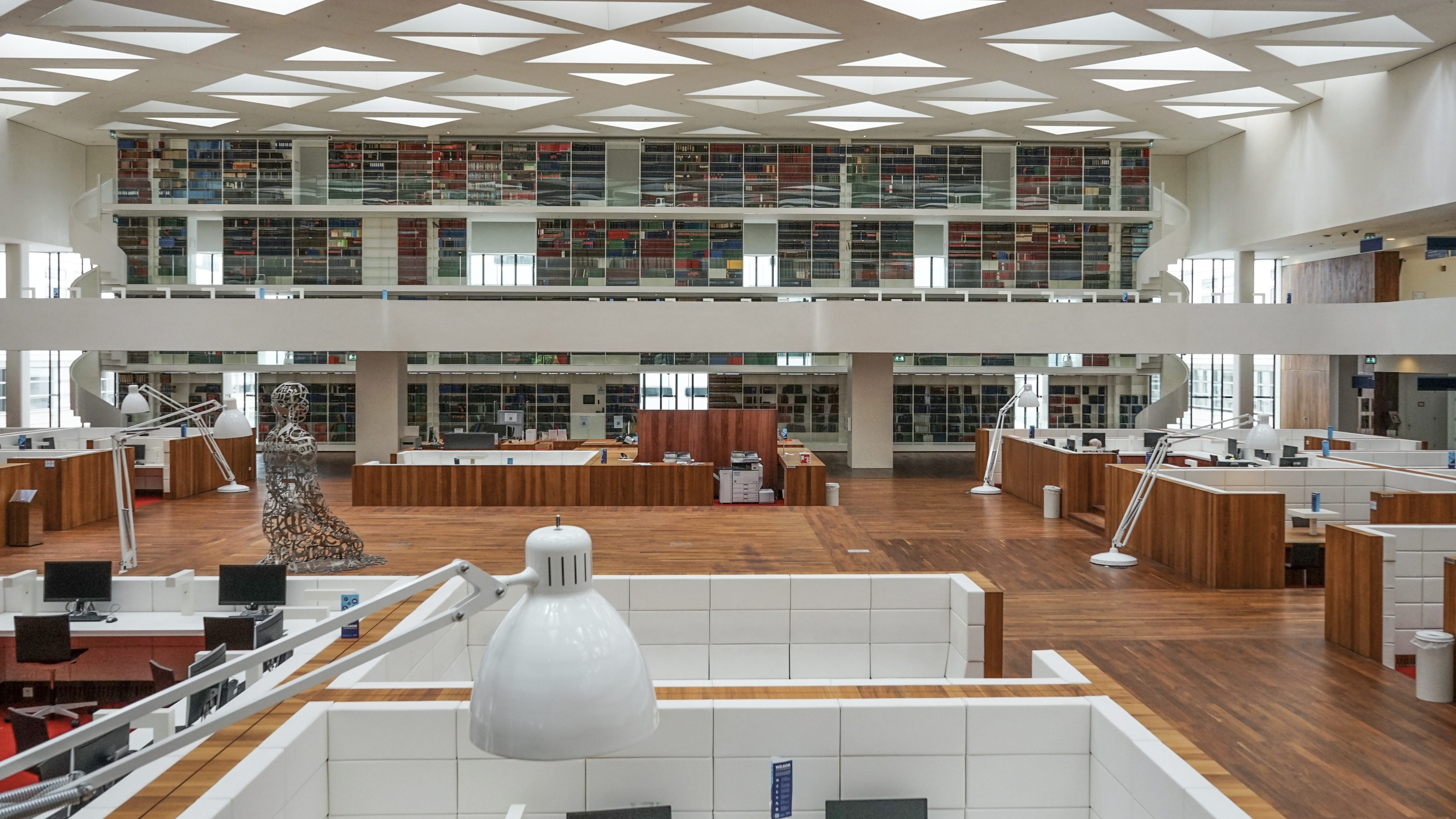 De moderne bibliotheekzaal valt op door geometrische daklichten die daglicht werpen op rijen werkplekken met monitoren. Afgeschermde individuele werkplekken, een grote metalen sculptuur als blikvanger en hoge boekenwanden bepalen het beeld. Een wenteltrap leidt naar boven - ideaal voor thema's als architectuur, onderwijs of geconcentreerd leren