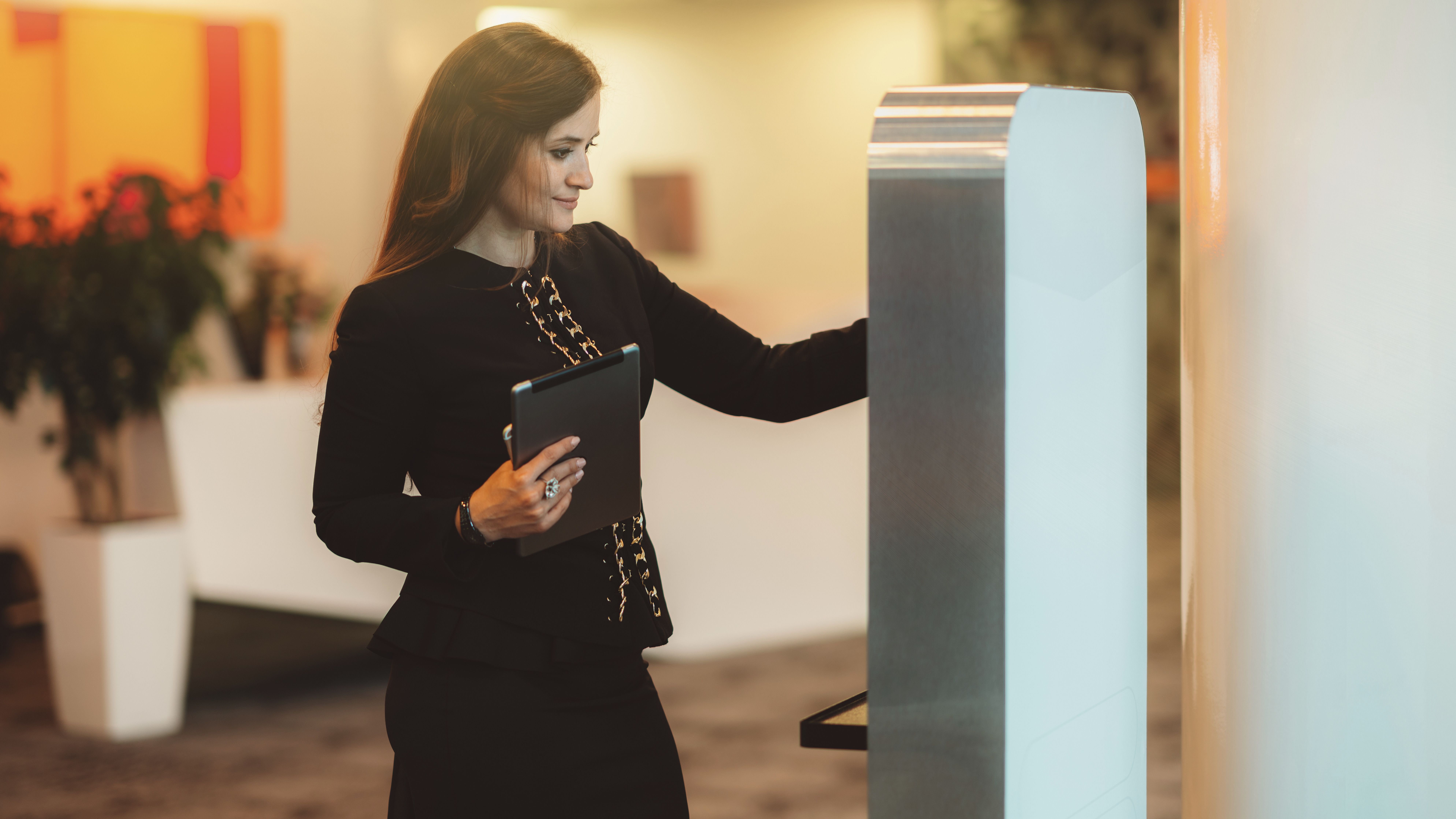Uma mulher de longo cabelo castanho está numa receção de hotel luminosa com um tablet na mão enquanto interage com um terminal. O seu traje de negócios preto sublinha o ambiente profissional.