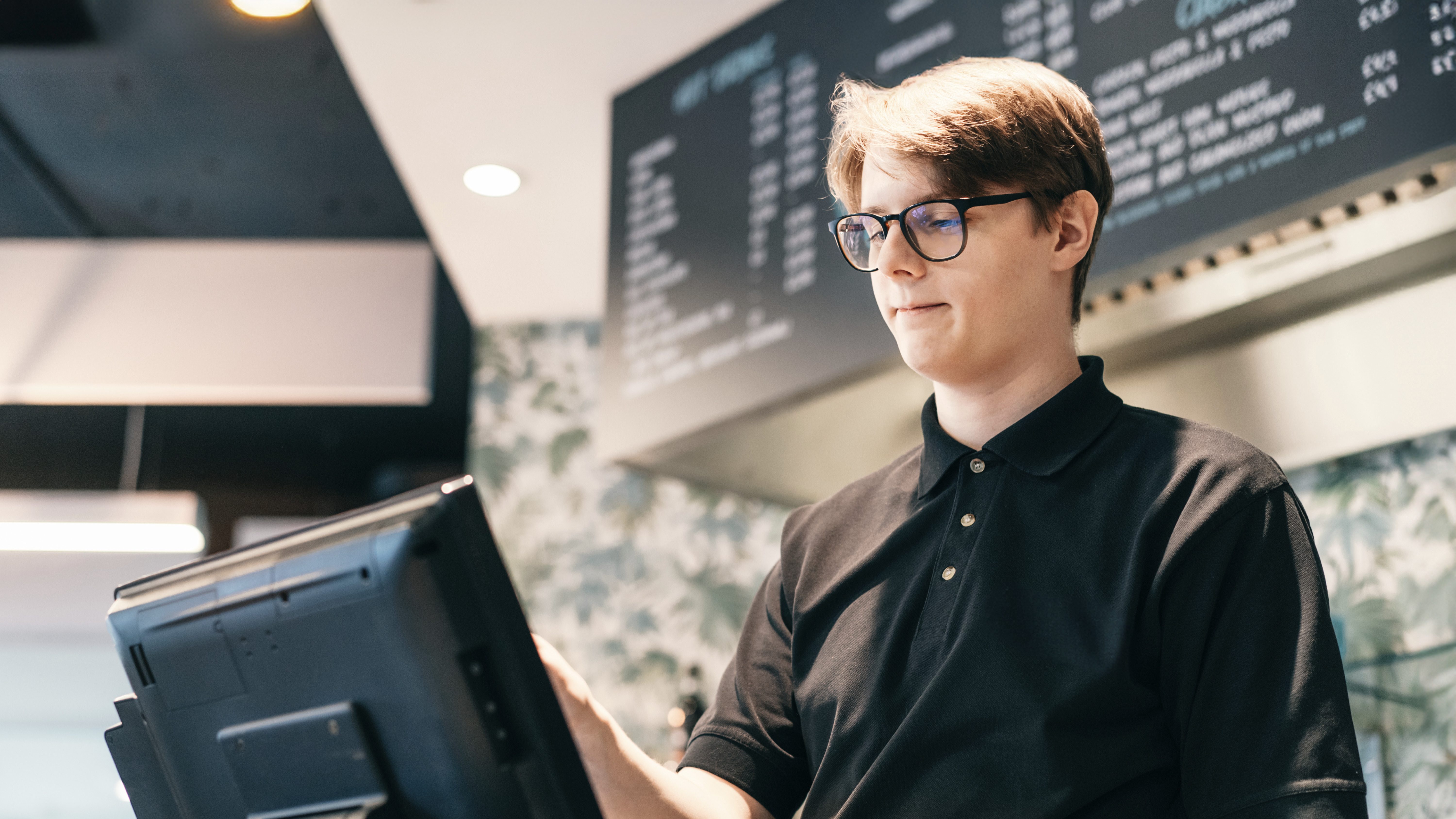 Ein junger Mann mit hellbraunem Haar und Brille arbeitet konzentriert an einem Touch-Monitor oder einer Kasse in einem hellen, modernen Arbeitsumfeld. Er trägt ein dunkles Poloshirt. Im Hintergrund befinden sich schwarze Displays mit einer Speisekarte in weißer Schrift.