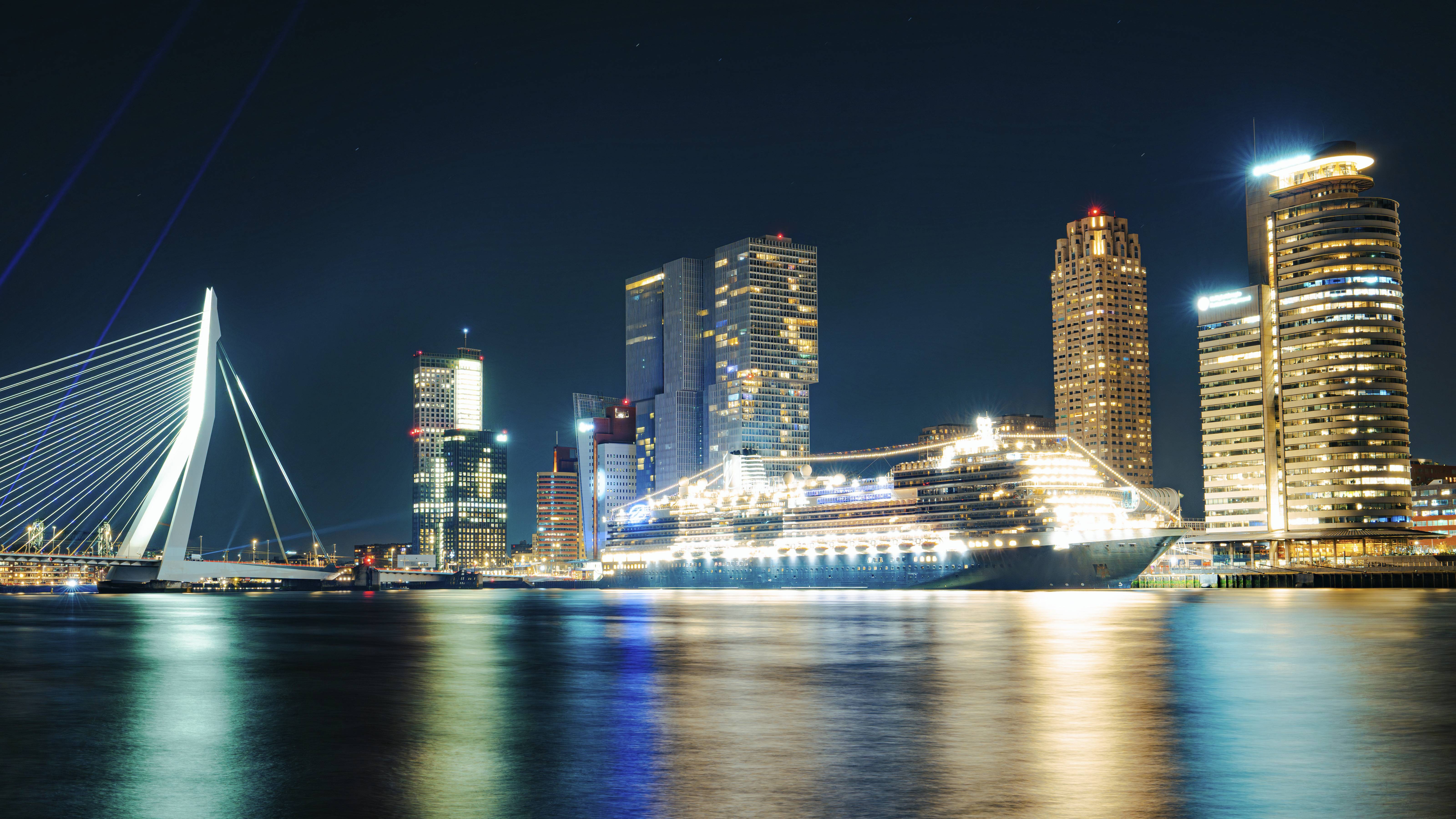 Une photo nocturne pittoresque montre la silhouette paisible d'une ville dont les lumières se reflètent dans l'eau. Au premier plan, un bateau de croisière illuminé brille de mille feux, symbole de voyages et d'aventures. L'ambiance harmonieuse éveille l'envie de voyager et convient parfaitement aux campagnes touristiques ou urbaines. Le pont à l'arrière-plan souligne le caractère urbain, tandis que les lumières sur l'eau sont idéales pour la promotion de croisières ou d'activités nocturnes.