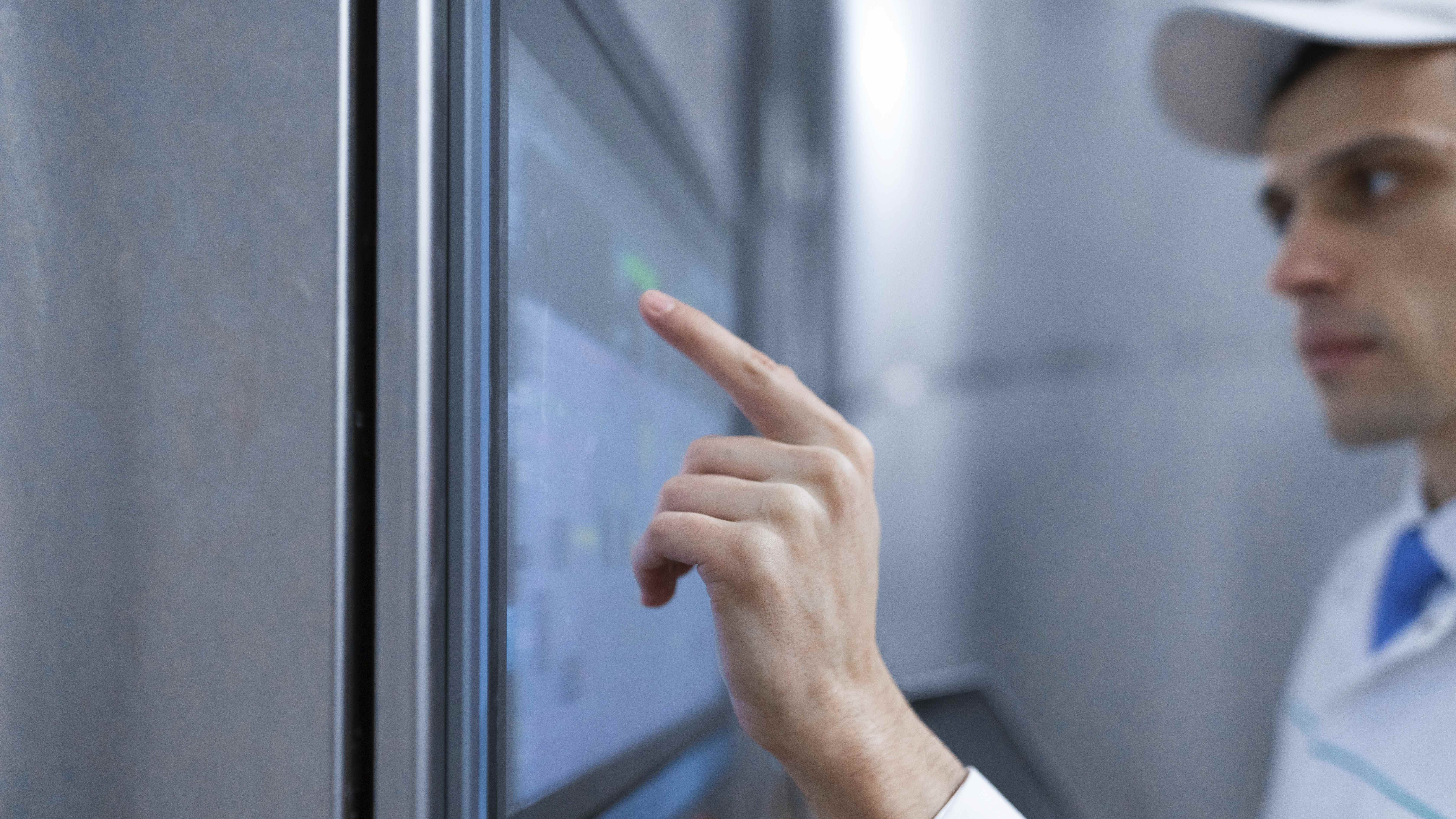 Ein Mann im weißen Kittel und mit Haube steht in einem hell erleuchteten Industrieumfeld und bedient einen Touchscreen. Metallwände und technische Geräte prägen die Szene – ein Bild für Projekte zu Technologie, Industrie, Automatisierung und modernen Arbeitswelten. Es vermittelt Fortschritt, Effizienz und Innovation.